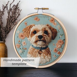 May include: Embroidered dog portrait in a wooden hoop. The dog has brown and white fur, a blue collar, and is surrounded by peach-coloured floral embroidery on a light blue background. The text "Handmade pattern template" is visible.
