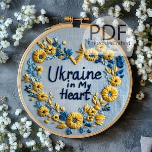 May include: A round embroidery hoop with the text "Ukraine in My Heart" in blue thread. The design features yellow and blue floral embroidery, with a wooden hoop and a light blue fabric background. The image includes the text "PDF" and "EmbroiderCrafted".