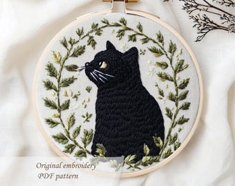 Puede incluir: Un bastidor de bordado con un diseño de gato negro. El gato está rodeado por una corona floral verde y blanca. El texto "Original embroidery PDF pattern" se muestra debajo.