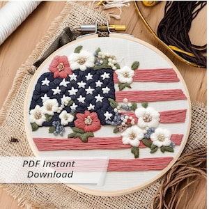 Pode incluir: Arte de bordado em aro com uma bandeira americana com flores brancas e listras vermelhas e brancas. O aro é de madeira e tem um acabamento natural. O texto "PDF Instant Download" é visível abaixo do aro.