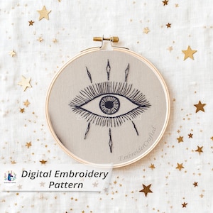 May include: A round embroidery hoop with a beige fabric featuring a dark blue embroidered eye design. The text "Digital Embroidery Pattern" is visible. Gold star-shaped confetti is scattered around the hoop.