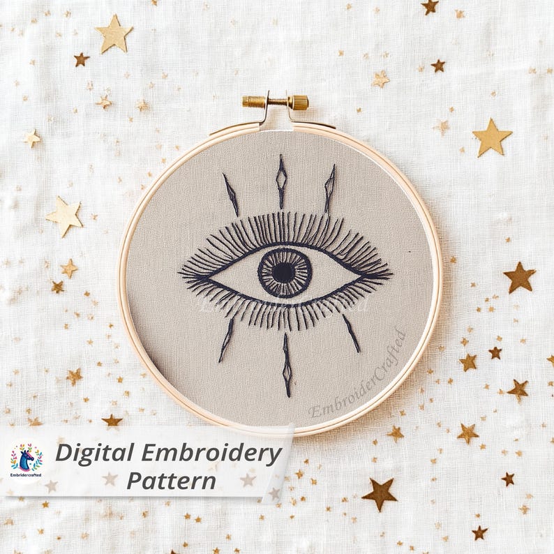 May include: A round embroidery hoop with a beige fabric featuring a dark blue embroidered eye design. The text "Digital Embroidery Pattern" is visible. Gold star-shaped confetti is scattered around the hoop.