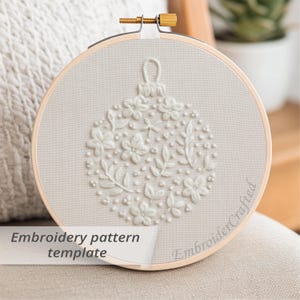 Puede incluir: Un bastidor de bordado crema con un diseño floral de adorno navideño. El diseño está en relieve con flores, hojas y puntos. El texto "Embroidery pattern template" se muestra en la parte inferior izquierda.