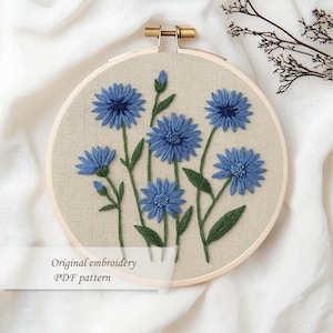 May include: Embroidered floral hoop art featuring blue flowers with green stems and leaves. The embroidery is set against a beige fabric background, framed by a wooden hoop. Text on the image reads "Original embroidery PDF pattern."