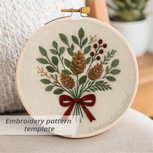 May include: An embroidery pattern template featuring a bouquet of pinecones, leaves, and berries, tied with a red bow. The design is stitched on a cream-colored fabric, held within a wooden embroidery hoop. The text "Embroidery pattern template" is visible.
