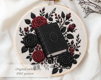 May include: Embroidered hoop art featuring a black book with ornate detailing, surrounded by red and black roses and foliage. The text "Original embroidery PDF pattern" is displayed below the design. The embroidery is set against a neutral fabric.