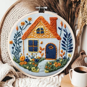 May include: Embroidered house in a wooden hoop. The house features an orange roof and chimney, blue door and windows, and a garden with colorful flowers. The embroidery is set against a light blue fabric background.