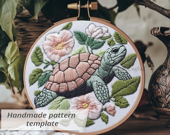 May include: Embroidered turtle design in a wooden hoop. The turtle has a brown shell and green skin, surrounded by pink flowers and green leaves. The text "Handmade pattern template" is displayed on the bottom left.