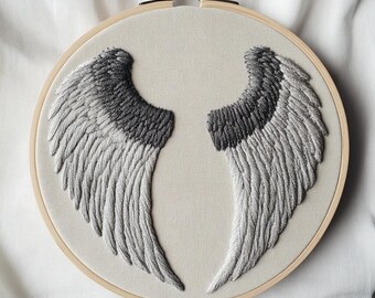 May include: An embroidery hoop features a detailed, hand-stitched design of two angel wings. The wings are rendered in shades of gray, with darker gray at the top and lighter gray towards the tips. The embroidery is set against a light beige fabric.