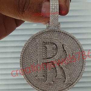 May include: A round, silver-colored pendant with the letter "R" in the center, encrusted with small, sparkling stones. The pendant is held by a hand and has a loop at the top for a chain.