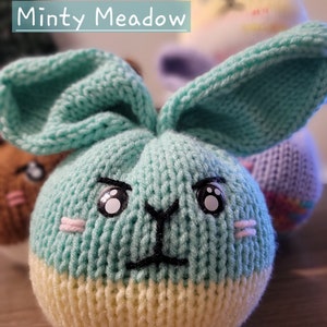 May include: A knitted green and yellow bunny with a black nose and large white eyes. The bunny is sitting on a wooden surface. The text "Minty Meadow" is visible above the bunny.