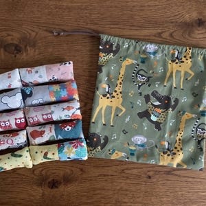 Può includere: Una collezione di oggetti in tessuto colorato su una superficie di legno. Ci sono diversi piccoli rotoli di tessuto fantasia e un sacchetto con coulisse con sfondo verde e una divertente stampa di giraffe e animali. Gli articoli sono probabilmente fatti a mano.