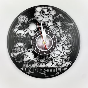 Può includere: Orologio in vinile bianco e nero con personaggi del videogioco Undertale. Il quadrante dell'orologio è un cerchio nero con numeri e lancette bianchi. I personaggi includono Sans, Papyrus, Flowey e Undyne.