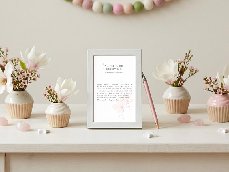 May include: A white picture frame containing a printed card titled A Letter to the Birthday Girl, with text inviting guests to write a message. A pink pen rests against the frame, flanked by four ceramic cupcake-shaped vases holding white flowers.