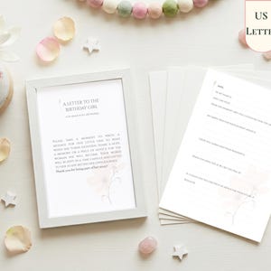 May include: A white frame containing a sign titled A LETTER TO THE BIRTHDAY GIRL, accompanied by several matching stationery cards with prompts for writing messages. The cards feature a soft floral design and are displayed with two pens.