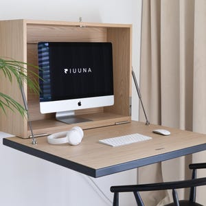 Peut inclure: Un bureau mural en bois clair avec une surface de travail rabattable soutenue par des tiges métalliques. L'intérieur du bureau contient un écran d'ordinateur avec le texte PIUUNA, un clavier blanc, une souris blanche et un casque audio blanc.