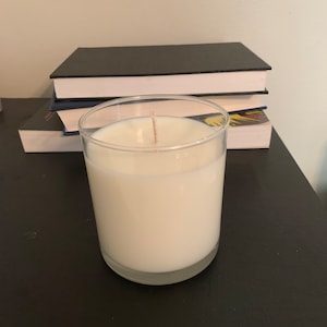 May include: A white candle in a clear glass jar with a single wick. The candle is sitting on a black surface with a stack of books behind it.