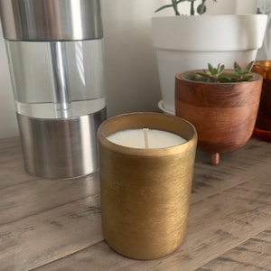 May include: A gold candle with a white wax top and a single wick. The candle is sitting on a wooden surface.