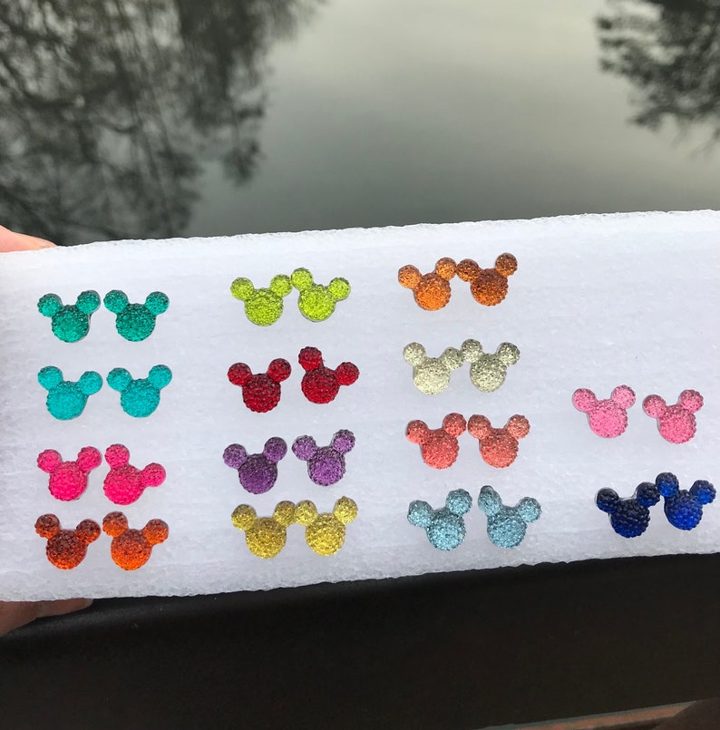 Puede incluir: Un tablero de espuma blanco con 12 pares de pendientes de colores en forma de cabeza de Mickey Mouse. Los pendientes est&aacute;n hechos de vidrio texturizado brillante. Los colores incluyen rojo, rosa, naranja, amarillo, verde, azul, morado y plateado.