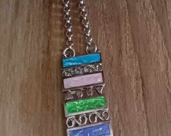 Puede incluir: Un collar de cadena de plata con un colgante de esmalte multicolor. El colgante está formado por cinco piezas rectangulares, cada una con un esmalte de color diferente. Los colores son azul, verde, rosa y turquesa.