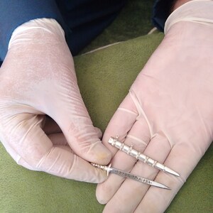 May include: Two silver-colored miniature swords, one with a decorative handle and the other with a plain handle, are held in gloved hands. The swords are about 4 inches long.