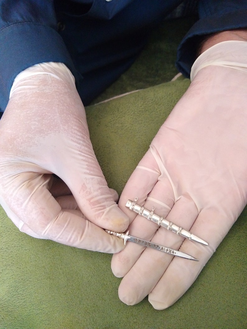 May include: Two silver-colored miniature swords, one with a decorative handle and the other with a plain handle, are held in gloved hands. The swords are about 4 inches long.