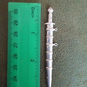 May include: A silver sword charm with intricate details, measuring approximately 2 inches in length.