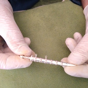 May include: A silver-colored, sword-shaped pendant with a pointed tip and two loops at the top. The pendant is about 5 inches long.