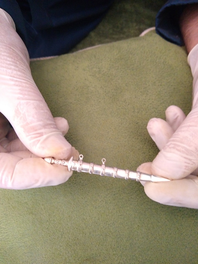 May include: A silver-colored, sword-shaped pendant with a pointed tip and two loops at the top. The pendant is about 5 inches long.