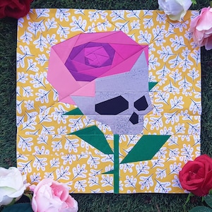 Puede incluir: Una flor de papel rosa y morada con una cabeza de calavera gris, sobre un fondo amarillo con un patrón de hojas blancas.