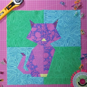 Puede incluir: Un bloque de colcha de gato rosa y morado con un patrón floral. El gato está sentado sobre un fondo verde y azul con un patrón de remolino.