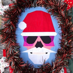 Puede incluir: Un bloque de colcha navideño festivo con la cara de Papá Noel con un gorro rojo, barba blanca y gafas de sol rosas. El bloque está rodeado de oropel rojo y dorado.