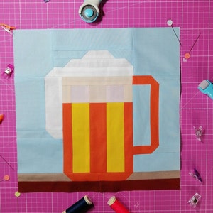 Puede incluir: Un bloque de colcha de retazos que representa una jarra de cerveza con una cabeza de espuma blanca, un cuerpo naranja y rayas amarillas. La jarra está sobre una base marrón sobre un fondo azul claro.