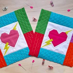 May include: Two unfinished quilt blocks with a white background, featuring a pink and red heart with a yellow lightning bolt design. The blocks are surrounded by turquoise, green, red, and orange fabric.