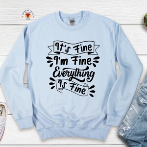 May include: Light blue sweatshirt with black text that reads "It's Fine I'm Fine Everything Is Fine" with a banner design.