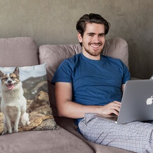 May include: A man sitting on a couch with a laptop on his lap. He is wearing a blue shirt and gray plaid pants. A throw pillow with a photo of a dog sitting on a rock is on the couch.