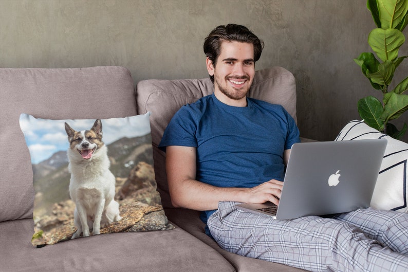 May include: A man sitting on a couch with a laptop on his lap. He is wearing a blue shirt and gray plaid pants. A throw pillow with a photo of a dog sitting on a rock is on the couch.