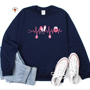May include: Navy blue sweatshirt with a pink Easter bunny and a pink heart with a stethoscope and a nurse's hat on it. The graphic is a heartbeat line with the bunny and heart in the middle.