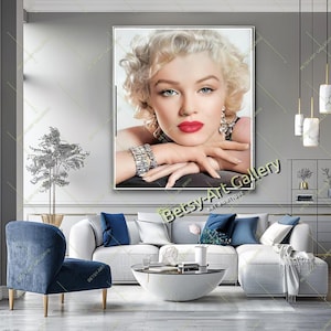 May include: A framed portrait of a woman with blonde hair, red lipstick, and blue eyes. She is wearing jewelry and resting her chin on her hands. The artwork is displayed above a white sofa with blue pillows.