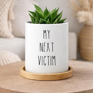 May include: A white cylindrical ceramic planter featuring the black text "MY NEXT VICTIM" printed on the front. The planter contains a green succulent plant and sits on a circular light brown wooden base, providing a stable foundation.