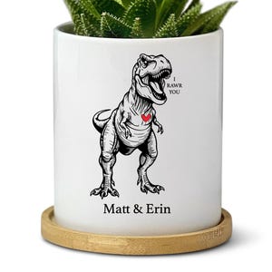May include: A white ceramic planter with a black and white illustration of a Tyrannosaurus Rex and a red heart. The text "I RAWR YOU" and "Matt & Erin" are also on the planter. The planter holds a green succulent plant and sits on a wooden coaster.
