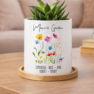 May include: A white ceramic planter with the words "Mom's Garden" and a colorful floral design. The planter holds a green succulent plant and sits on a round wooden coaster. The names Samantha, Niko, Ann, Robert, and Mandy are listed.