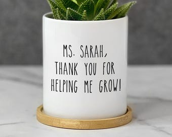 May include: A white cylindrical ceramic planter with a bamboo saucer base containing a green succulent plant. The front of the planter features black printed text that reads: MS. SARAH, THANK YOU FOR HELPING ME GROW! The design is simple and clean.