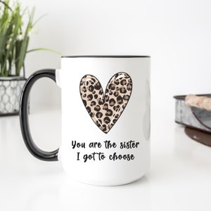 Op de afbeelding: Witte keramische mok met een zwarte rand en handvat. De mok heeft een hart met luipaardprint en de tekst "You are the sister I got to choose".