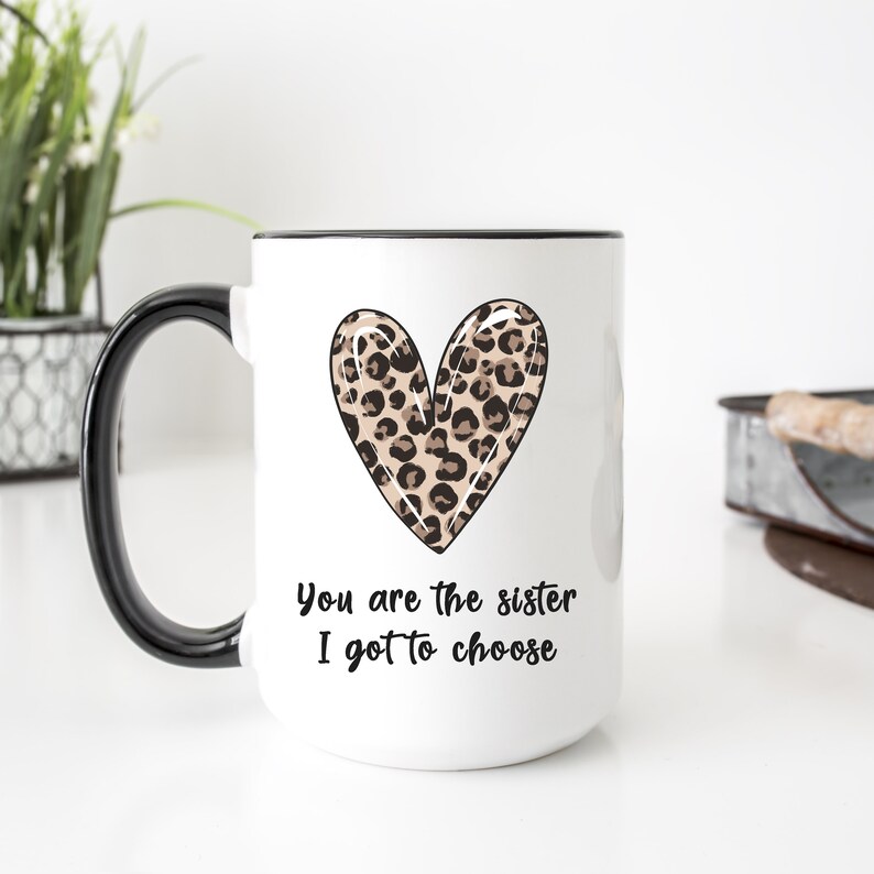 Op de afbeelding: Witte keramische mok met een zwarte rand en handvat. De mok heeft een hart met luipaardprint en de tekst "You are the sister I got to choose".