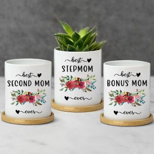 May include: Three white ceramic cylindrical planters, each resting on a circular bamboo coaster. The planters feature floral graphics with red roses and green leaves, along with the black text "best SECOND MOM ever", "best STEPMOM ever", and "best BONUS MOM ever".