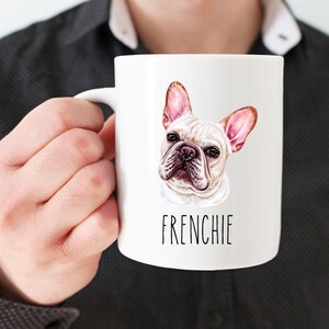 May include: White ceramic mug with a French Bulldog illustration, showing pink-lined ears and the word "FRENCHIE". The mug is held by a person wearing a black shirt.