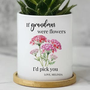 May include: A white cylindrical ceramic planter sitting on a circular bamboo tray. The planter features a watercolor illustration of pink flowers and black text that reads: If grandmas were flowers I'd pick you. LOVE, MELINDA. A green succulent plant is visible.