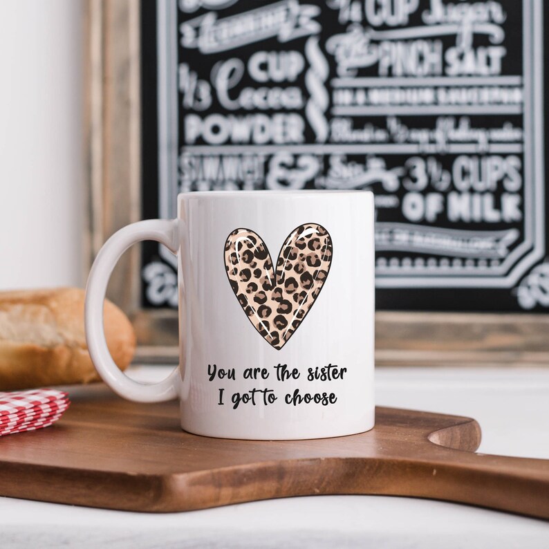 Op de afbeelding: Witte keramische mok met een luipaardprint hartontwerp en de tekst "You are the sister I got to choose".