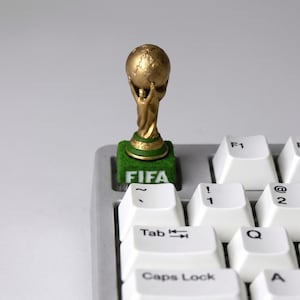 May include: A custom mechanical keyboard keycap featuring a miniature gold-colored FIFA World Cup trophy replica mounted on a green, textured square base with the white text FIFA printed on the front, positioned on a silver keyboard.